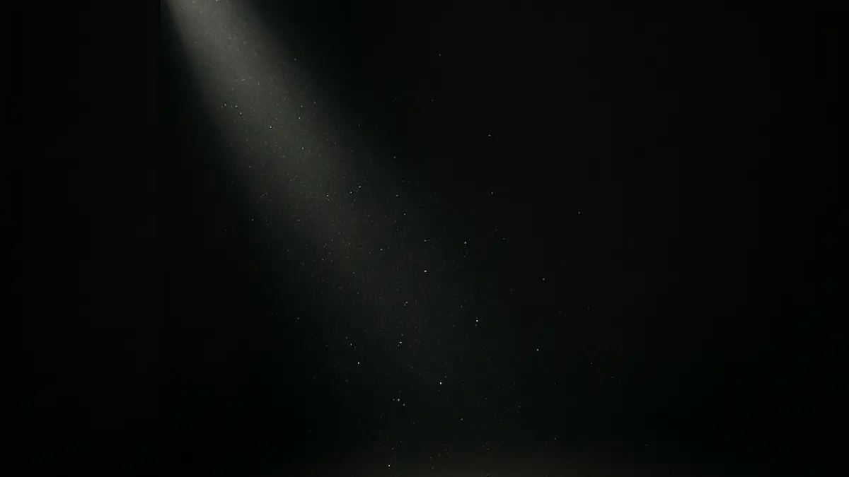 Ein einzelner Lichtstrahl im dunklen Raum, voll schwebender Staubpartikel. · A lone beam of light in a dark room, filled with drifting dust.